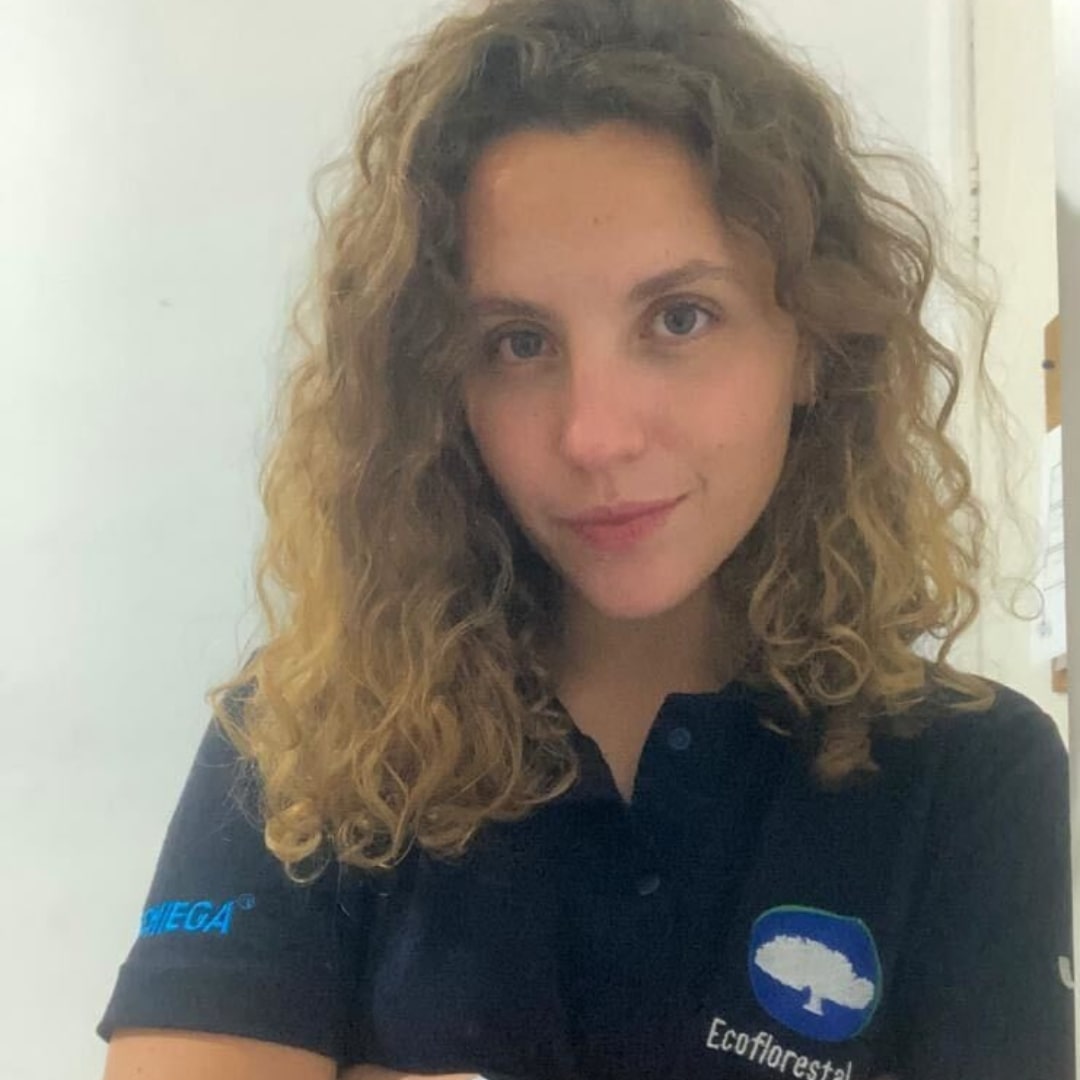 Descrição da imagem da Ana: pele clara, olhos escuros, cabelo médio, cacheado e loiro, está usando uma camisa polo azul escuro com o logo da EcoFlorestal e está sorrindo levemente pra foto