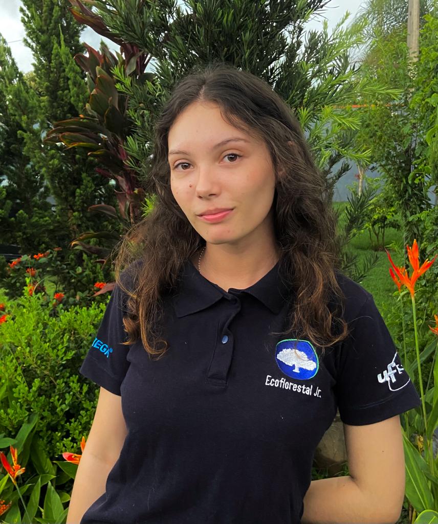 Descrição da imagem da Munique: pele clara, olhos escuros, cabelo castanho, está usando uma camisa polo azul escuro com o logo da EcoFlorestal e está sorrindo levemente pra foto